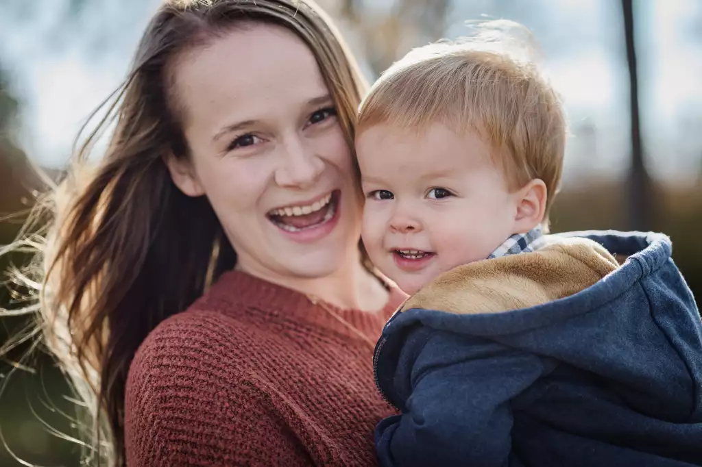 : Close-up of a smiling woman with long brown hair wearing a rust-colored sweater, holding a young child with light hair in a blue jacket with a beige lining, both looking towards the camera outdoors.