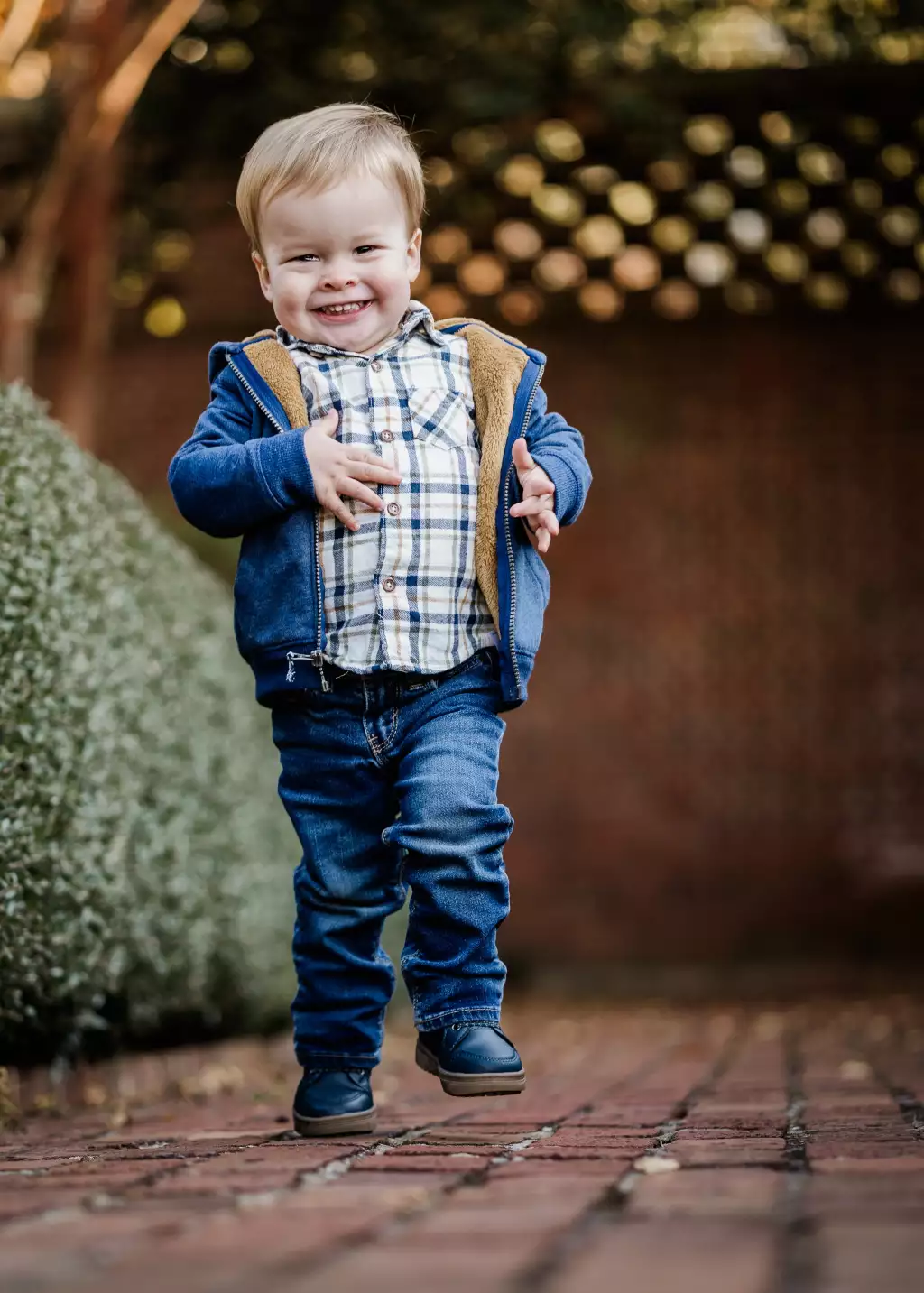 : Smiling toddler with blonde hair wearing a blue jacket with brown lining, plaid shirt, blue jeans, and dark shoes, walking on a brick pathway with green shrubbery on the left and a blurred background of lights and foliage.