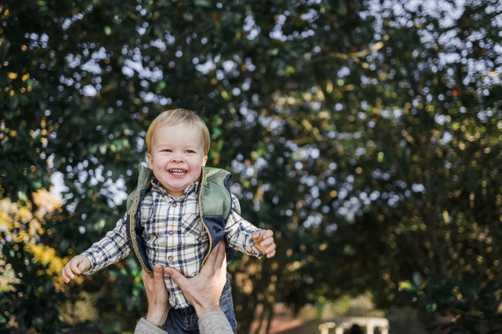 : Smiling toddler in a plaid shirt and green vest being lifted up outdoors by adult hands, with blurred green foliage in the background.