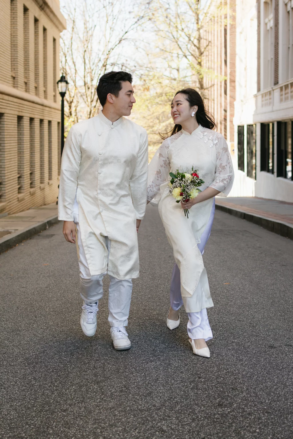 Robby and Hannah's couple photo session taken Downtown, Greenville: Couple in traditional white Vietnamese áo dài walking hand in hand down a city street, with the woman holding a bouquet of flowers.