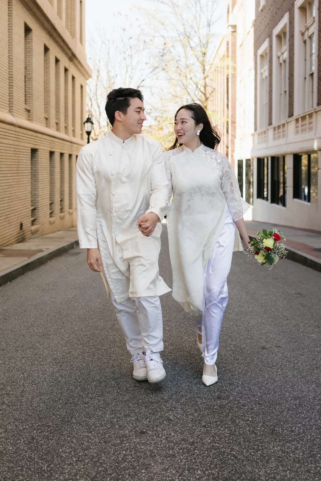 Robby and Hannah's couple photo session taken Downtown, Greenville: Couple in traditional white attire holding hands and smiling at each other while walking down a city street, woman holding a bouquet of flowers.