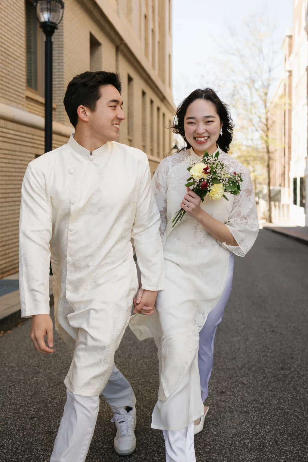 Robby and Hannah's couple photo session taken Downtown, Greenville: Smiling couple dressed in traditional white Vietnamese áo dài, holding hands and walking on a city street, woman holding a small bouquet of flowers.