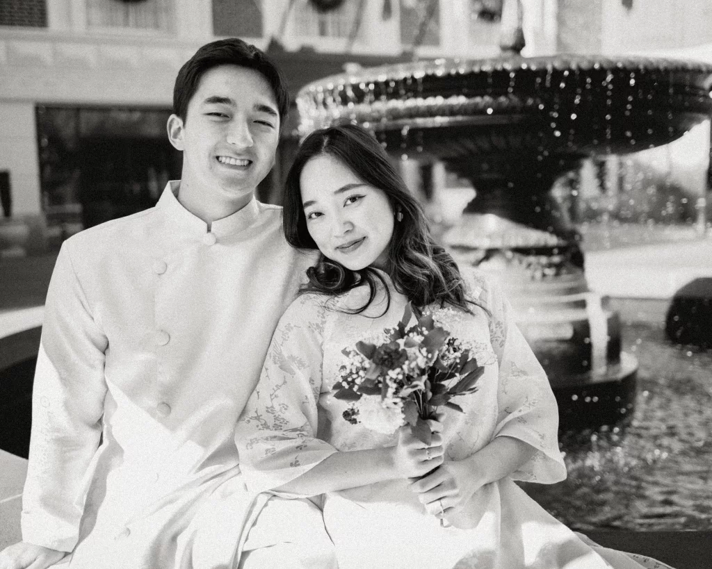 Robby and Hannah's couple photo session taken Downtown, Greenville: Black and white photo of a smiling couple dressed in traditional attire, the woman holding a bouquet, sitting by a fountain.