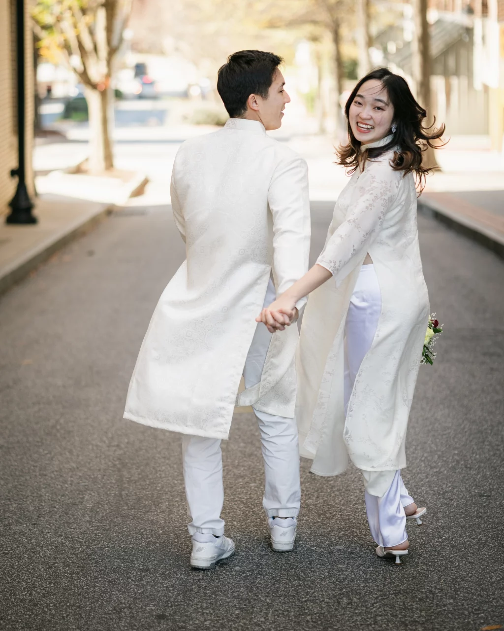 Robby and Hannah's couple photo session taken Downtown, Greenville: Couple in white traditional attire holding hands and walking away on a street, woman looking back and smiling.