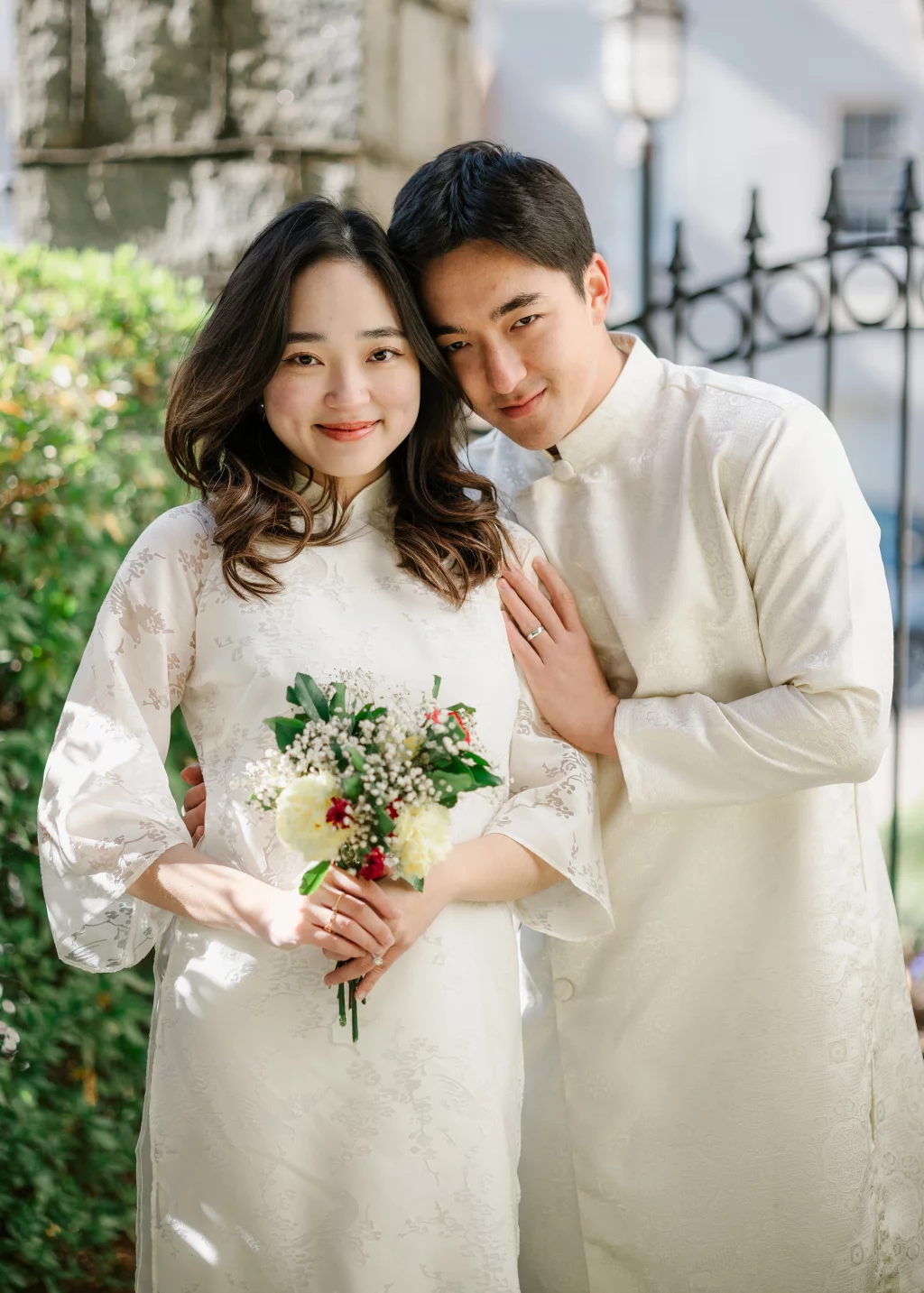 Robby and Hannah's couple photo session taken Downtown, Greenville: Portrait of a smiling couple in traditional white attire, with the woman holding a bouquet of white and red flowers, and the man leaning gently on her shoulder.