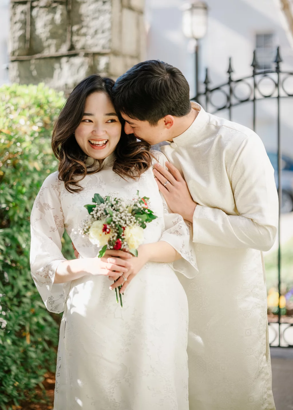 Robby and Hannah's couple photo session taken Downtown, Greenville: Bride and groom in traditional white attire, bride holding bouquet, groom leaning in close and smiling outdoors.