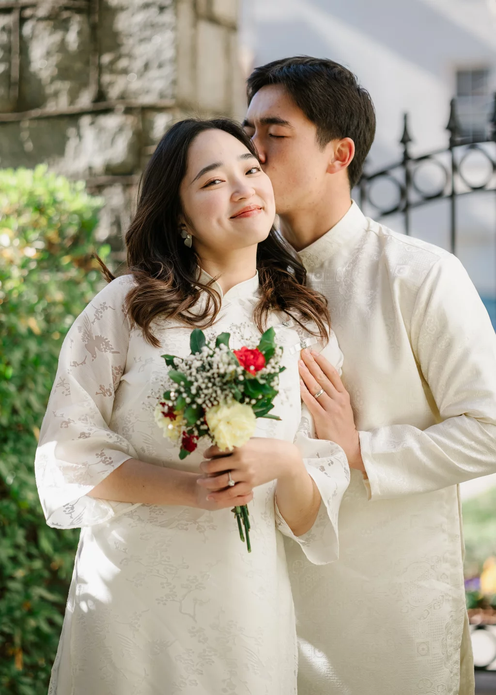 Robby and Hannah's couple photo session taken Downtown, Greenville: Bride holding a bouquet of flowers smiling as groom kisses her cheek, both dressed in traditional white attire.