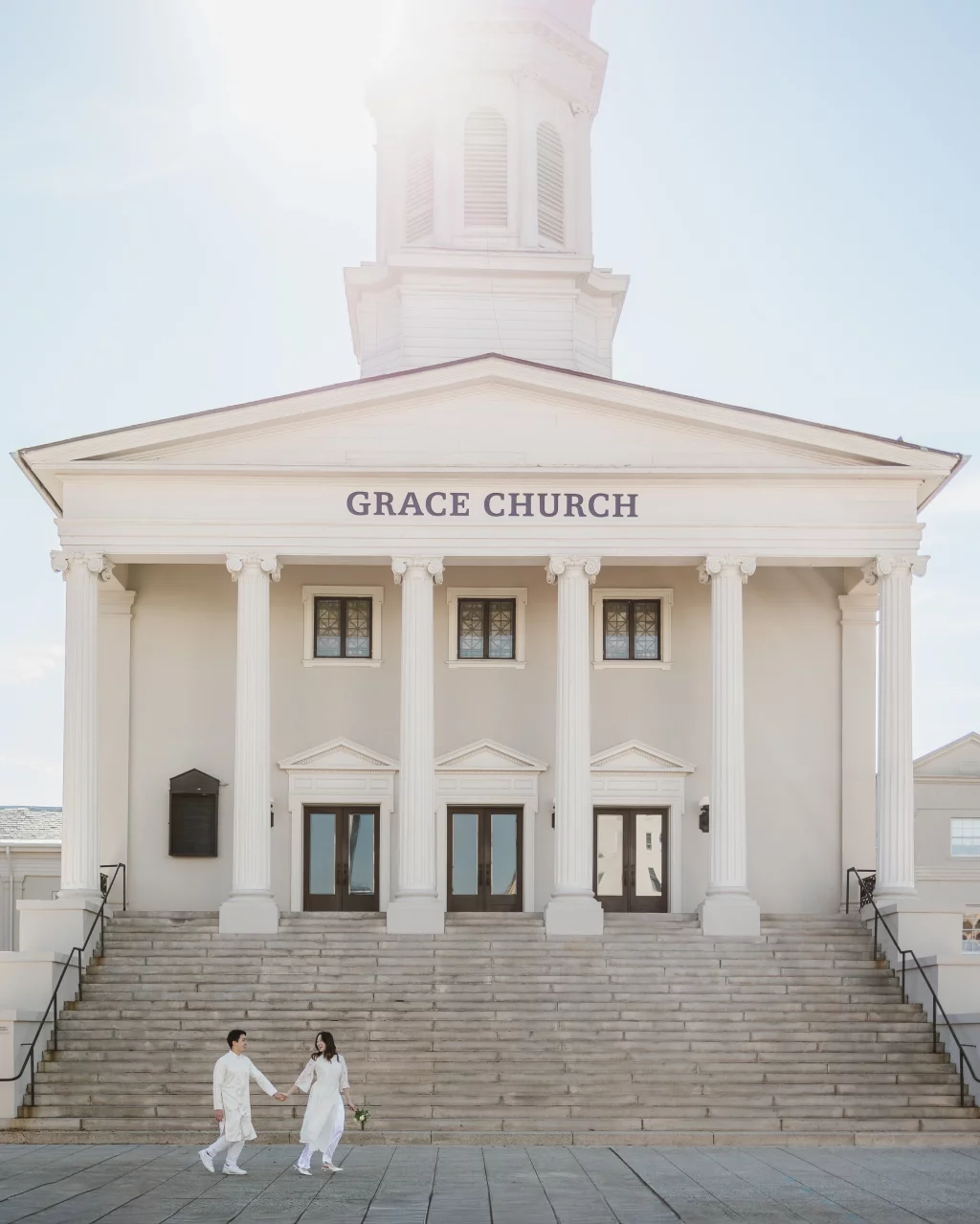 Robby and Hannah's couple photo session taken Downtown, Greenville: Front view of Grace Church with white columns and wide stone steps; a couple dressed in white holding hands at the bottom of the steps.