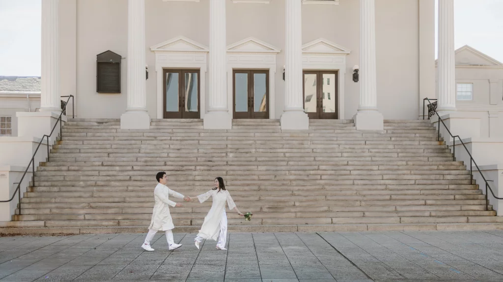 Robby and Hannah's couple photo session taken Downtown, Greenville: Couple in traditional white attire holding hands and walking in front of large stone steps and a building with three doors and tall columns.