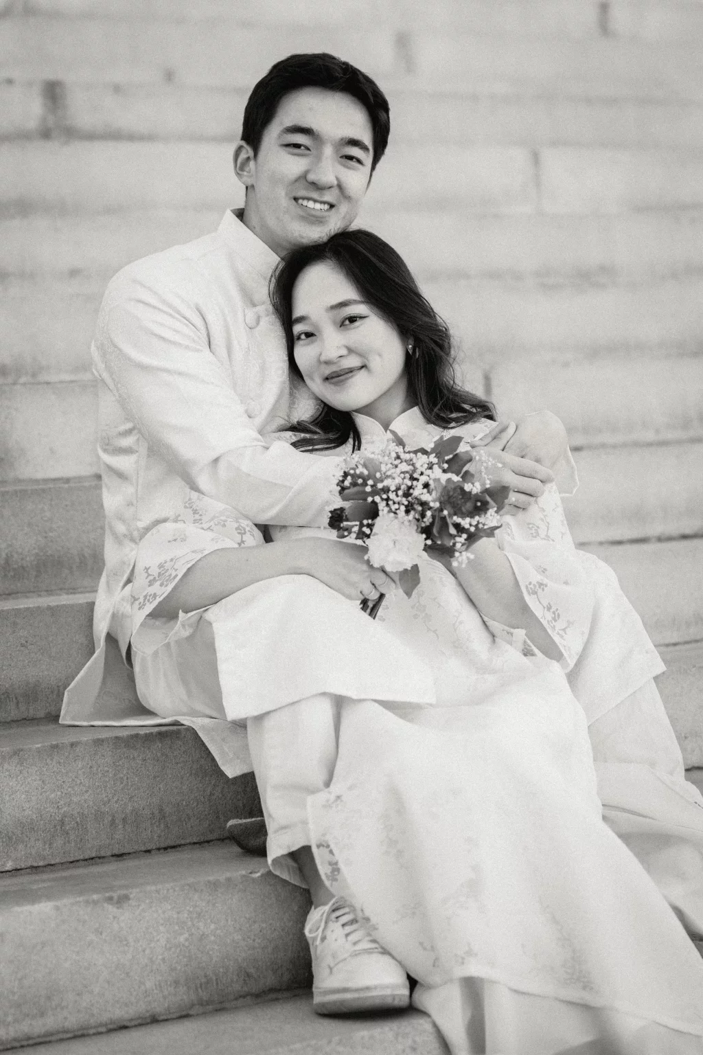 Robby and Hannah's couple photo session taken Downtown, Greenville: Black and white photo of a couple sitting on stone steps, the man embracing the woman who is holding a bouquet of flowers, both smiling.