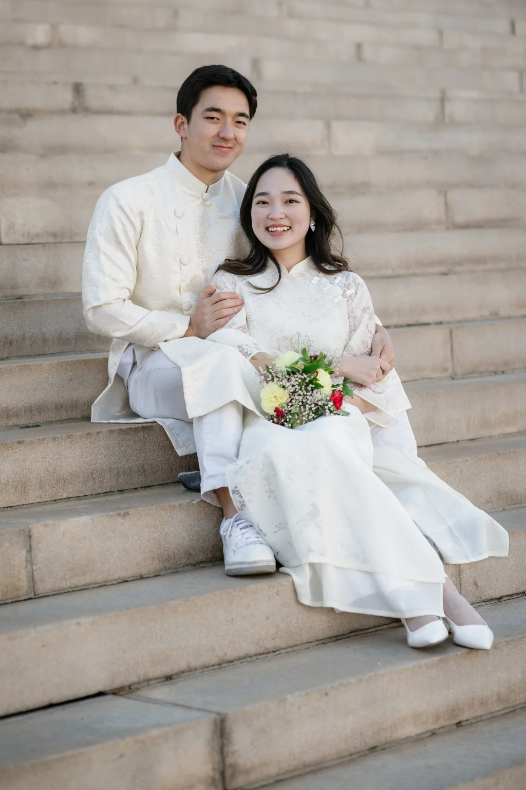 Robby and Hannah's couple photo session taken Downtown, Greenville: Young couple in traditional white attire sitting on stone steps, woman holding a bouquet of flowers and smiling at the camera.