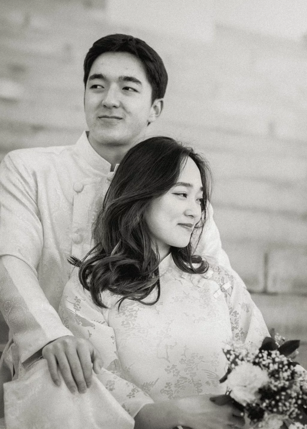 Robby and Hannah's couple photo session taken Downtown, Greenville: Black and white photo of a couple in traditional attire, the man standing behind the woman who is holding a bouquet of flowers, both looking slightly away from the camera.