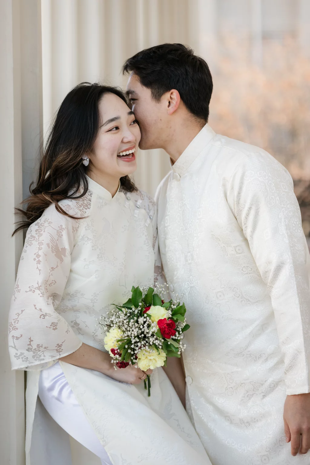 Robby and Hannah's couple photo session taken Downtown, Greenville: Young couple in traditional white Vietnamese áo dài, man whispering to smiling woman holding a bouquet of yellow and red flowers with greenery.