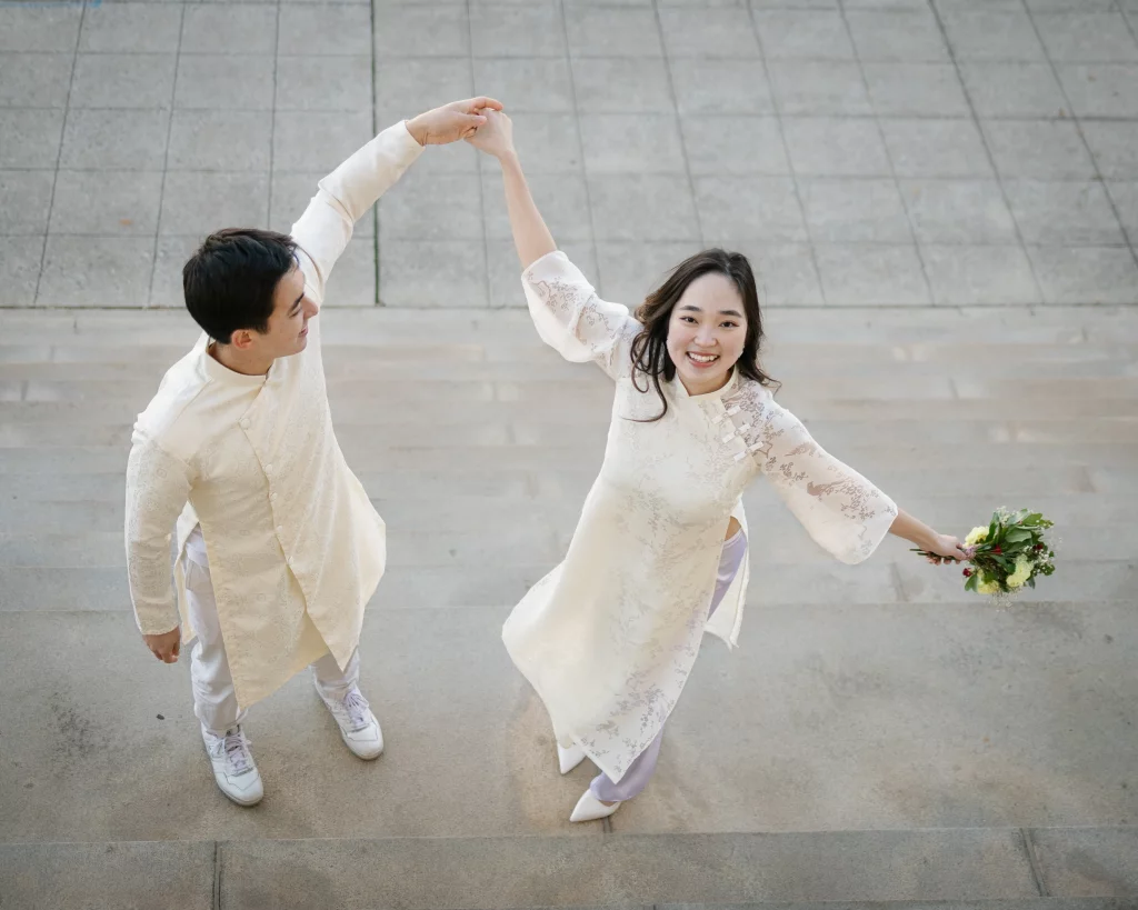 Robby and Hannah's couple photo session taken Downtown, Greenville: Overhead view of a smiling couple holding hands, dressed in traditional light-colored attire, with the woman holding a bouquet of flowers and standing on steps.