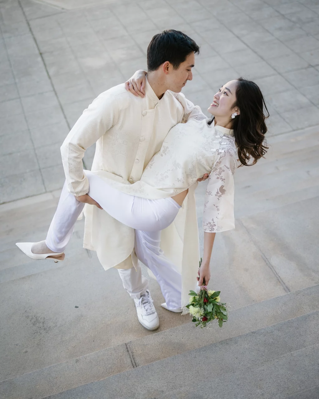 Robby and Hannah's couple photo session taken Downtown, Greenville: Man in traditional light-colored attire carrying a smiling woman in matching outfit holding a bouquet on outdoor steps.
