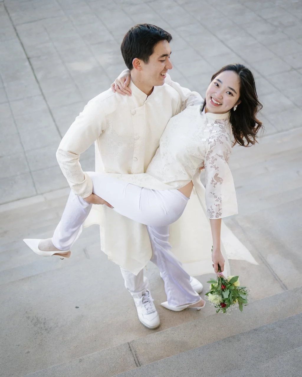 Robby and Hannah's couple photo session taken Downtown, Greenville: Man in traditional cream outfit holding and smiling at a woman in matching attire with a bouquet, standing on stone steps.