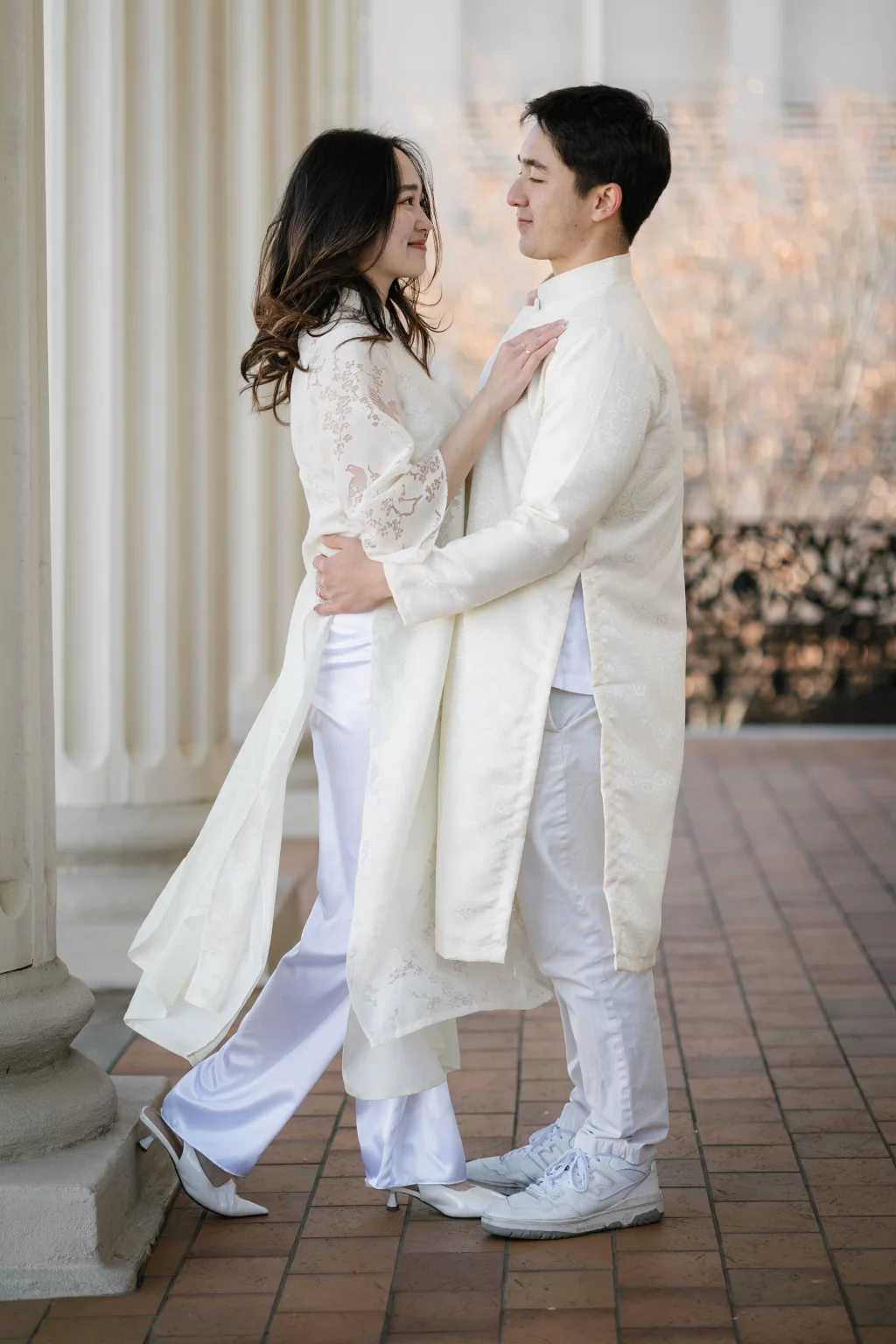 Robby and Hannah's couple photo session taken Downtown, Greenville: Couple in traditional white Vietnamese Ao Dai embracing and smiling at each other on a tiled floor with columns in the background.
