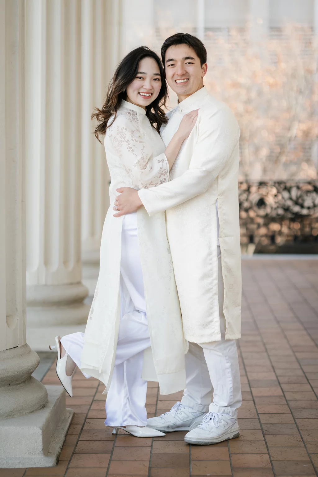 Robby and Hannah's couple photo session taken Downtown, Greenville: Portrait of a smiling couple in traditional white attire standing close and embracing near white columns on a tiled floor.
