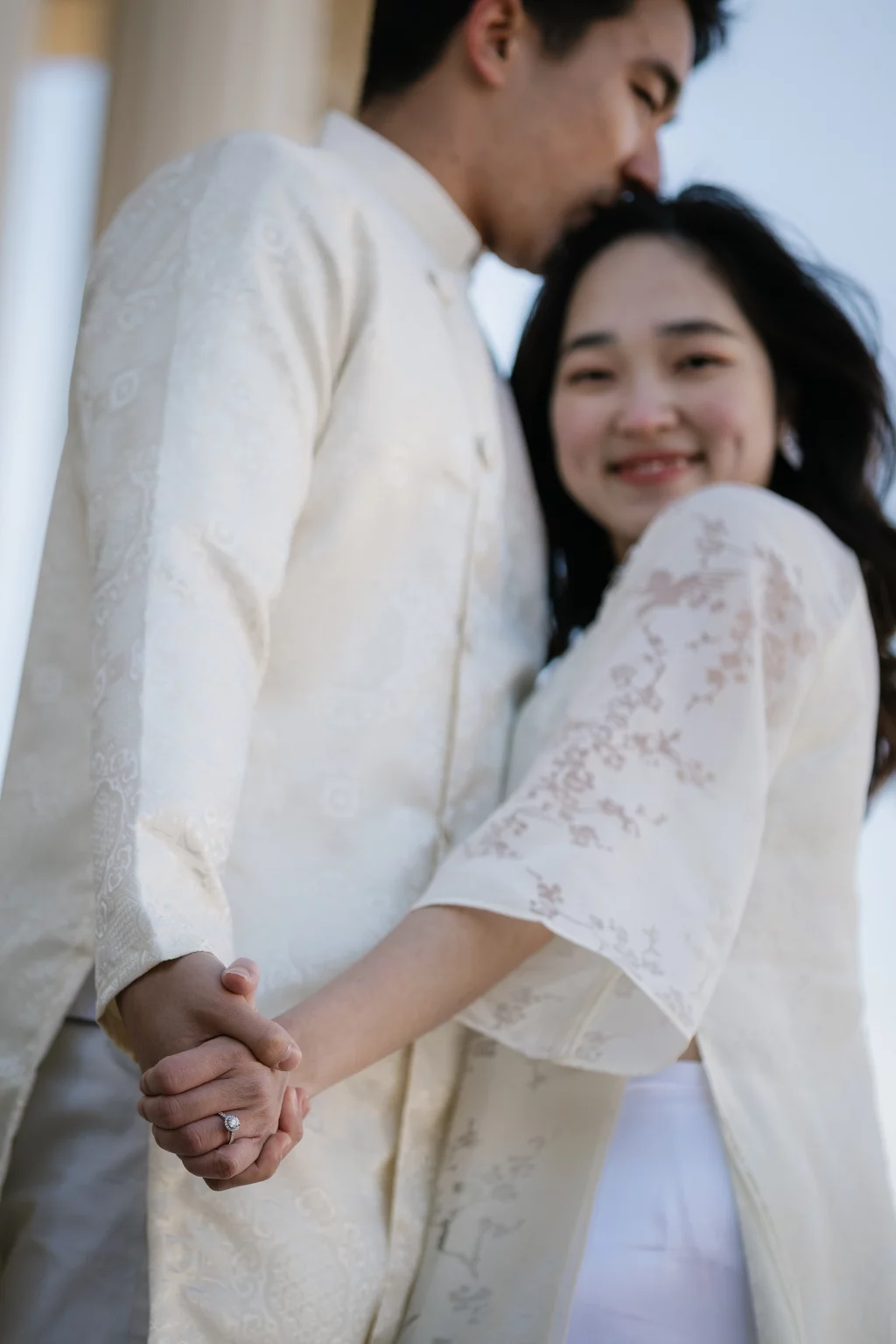 Robby and Hannah's couple photo session taken Downtown, Greenville: Close-up of a couple dressed in traditional white attire holding hands, the woman smiling with an engagement ring visible, and the man kissing her forehead.