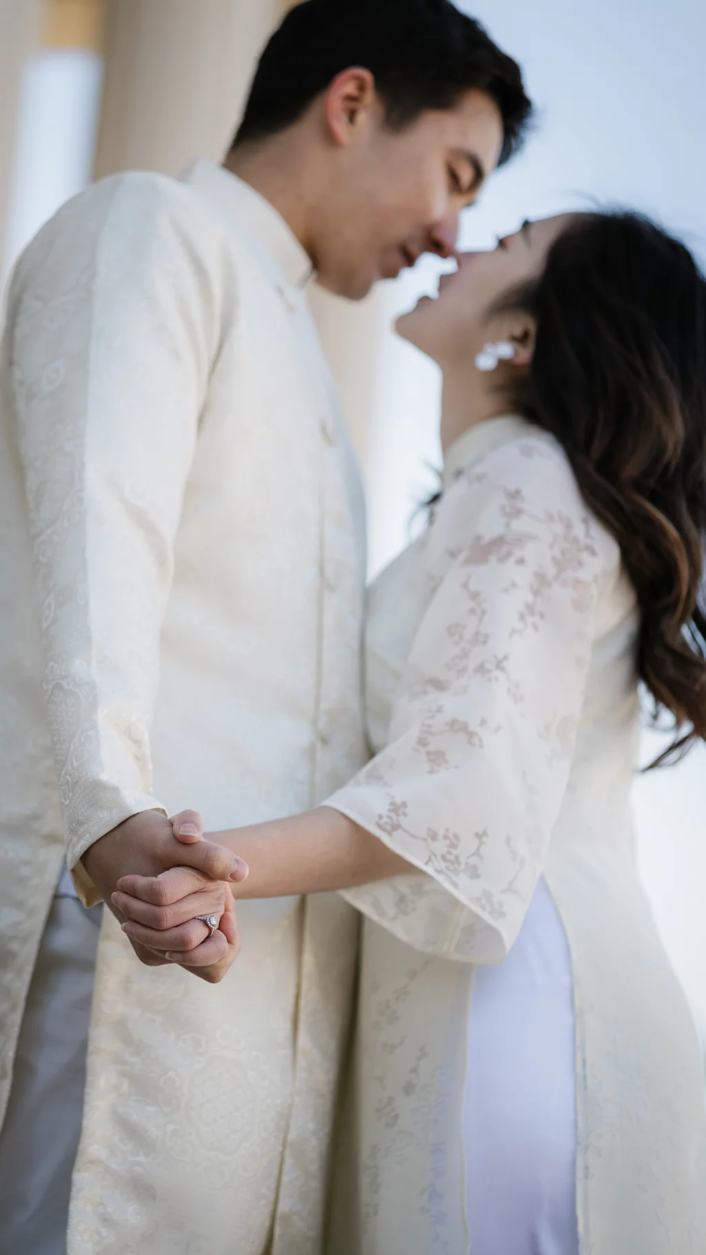 Robby and Hannah's couple photo session taken Downtown, Greenville: Close-up of a couple holding hands and about to kiss, both wearing traditional light-colored embroidered outfits, with a focus on the woman's engagement ring.