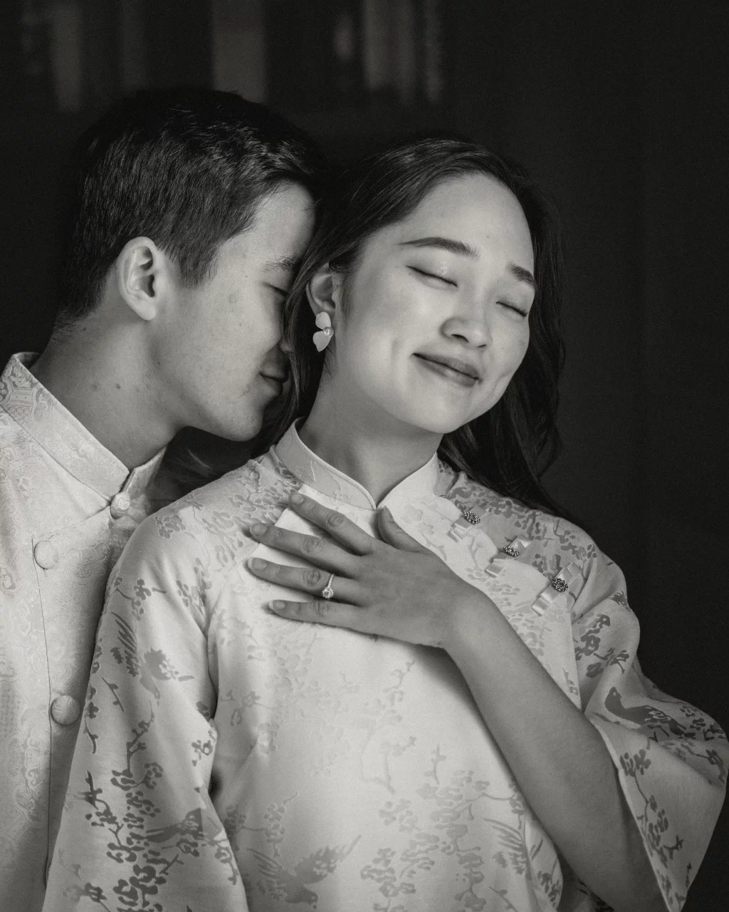 Robby and Hannah's couple photo session taken Downtown, Greenville: Black and white photo of a couple in traditional attire, the man gently nuzzling the woman's ear as she smiles with eyes closed, her hand displaying an engagement ring.