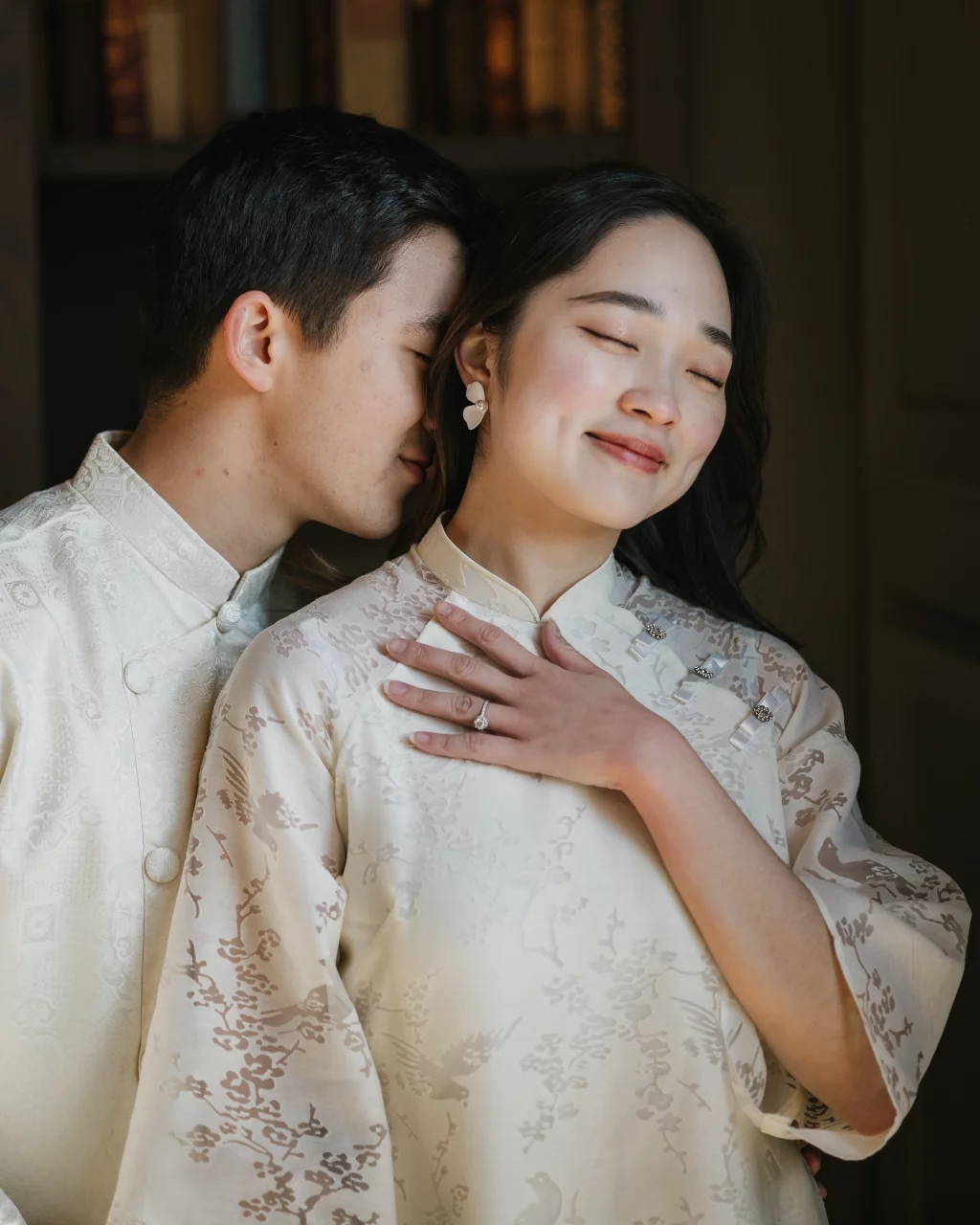 Robby and Hannah's couple photo session taken Downtown, Greenville: A couple dressed in matching light cream traditional attire, the man gently kissing the woman's cheek as she smiles with eyes closed and hand over her chest, showing an engagement ring.