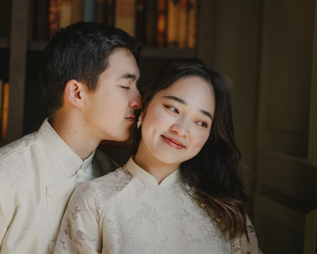: Portrait of a couple in traditional matching light-colored attire; the man is gently kissing the woman on the cheek while she smiles softly.