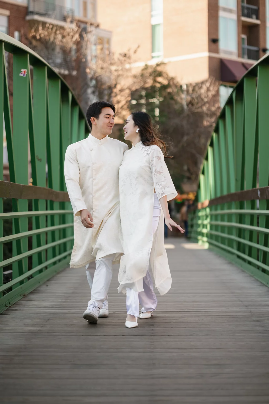 Robby and Hannah's couple photo session taken Downtown, Greenville: Young couple wearing traditional white Vietnamese ao dai walking arm in arm on a green pedestrian bridge.