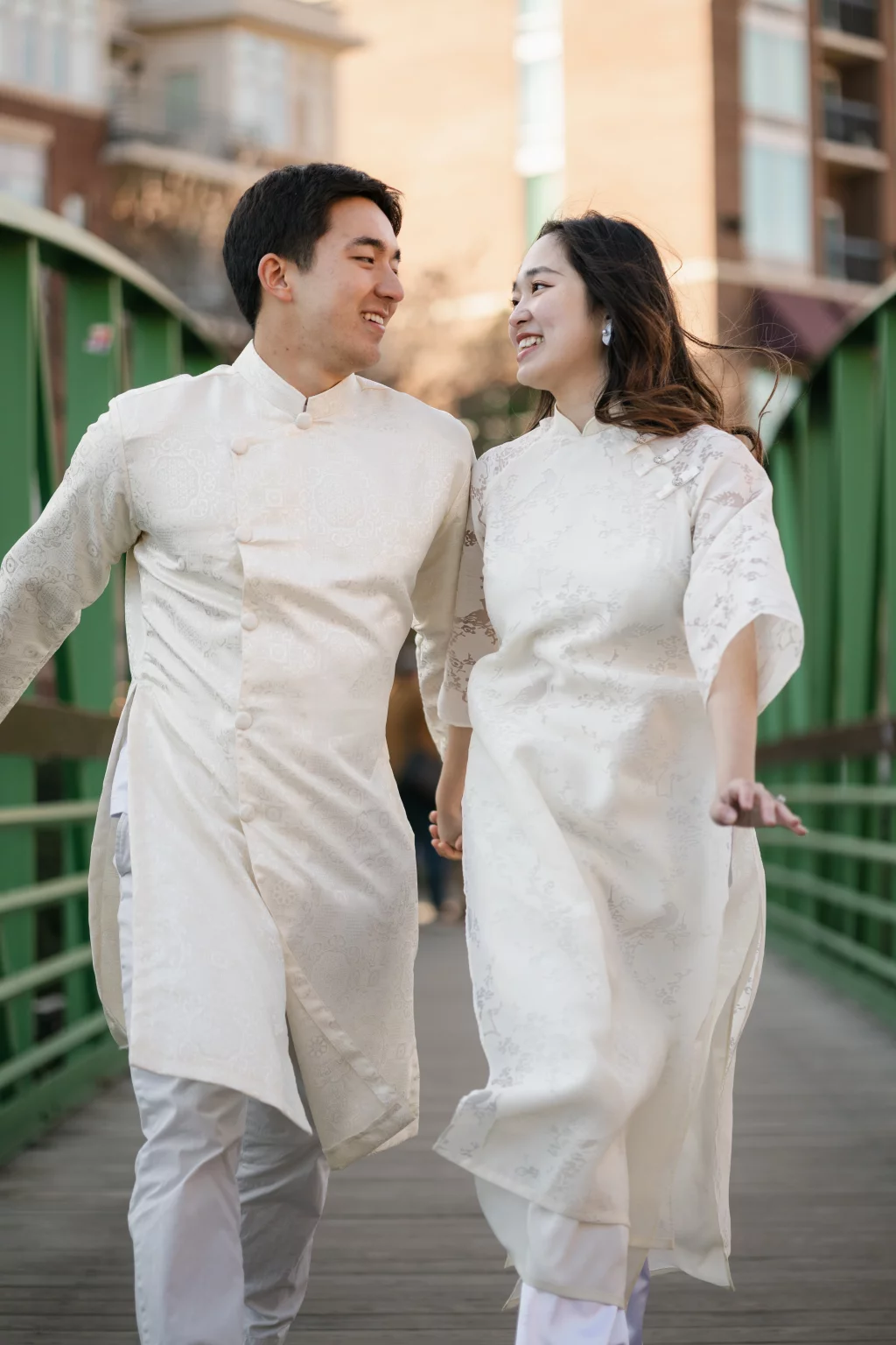 Robby and Hannah's couple photo session taken Downtown, Greenville: Couple in traditional matching white Vietnamese áo dài walking and smiling at each other on a green bridge.