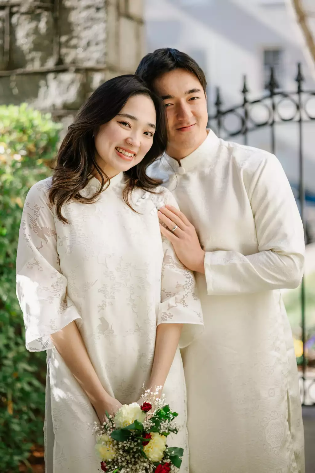 : Smiling couple dressed in matching traditional white attire, woman holding a bouquet of flowers, standing outdoors near a stone wall and wrought iron fence.