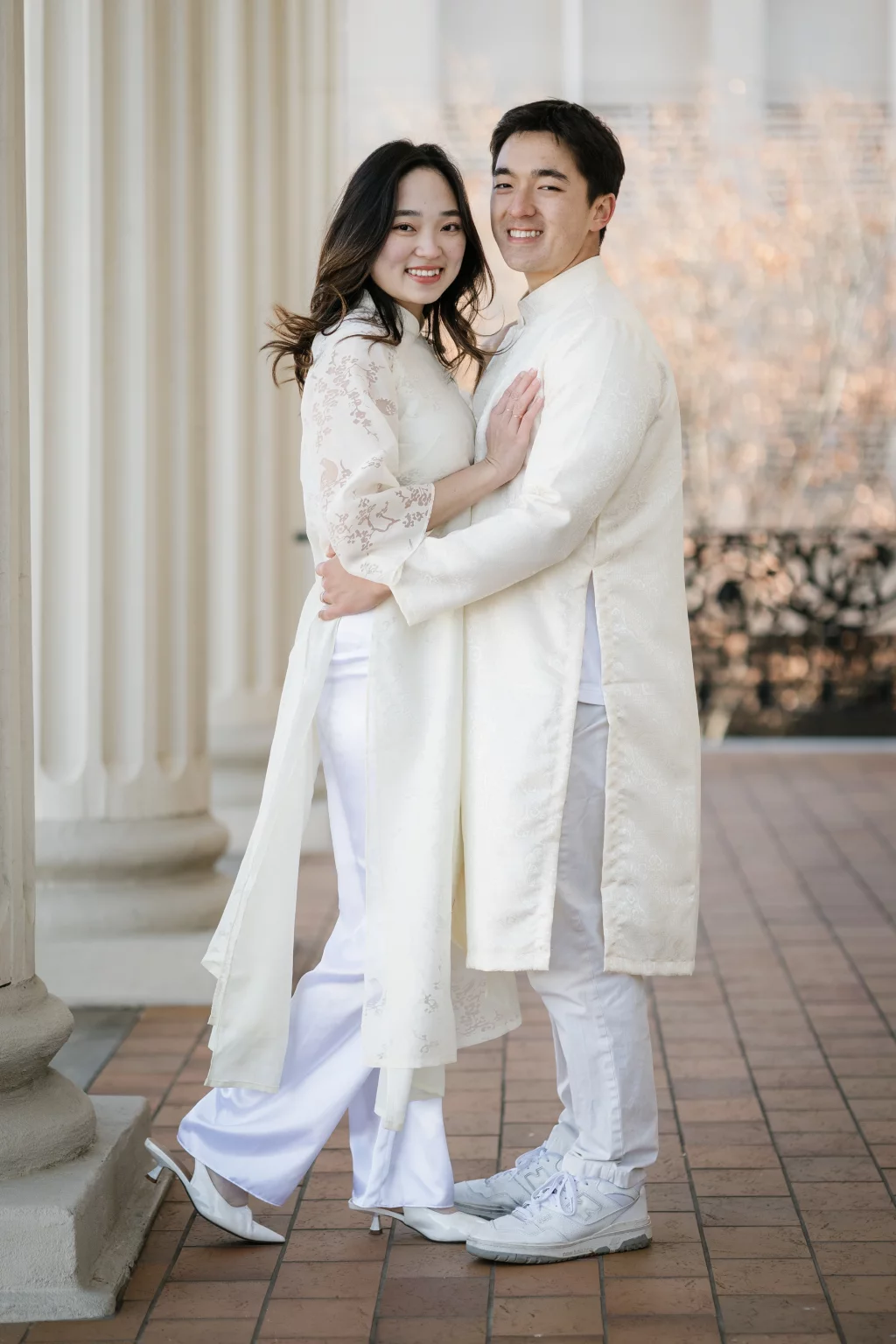 Robby and Hannah's couple photo session taken Downtown, Greenville: Couple in matching white traditional outfits embracing and smiling, standing on tiled floor by large columns.