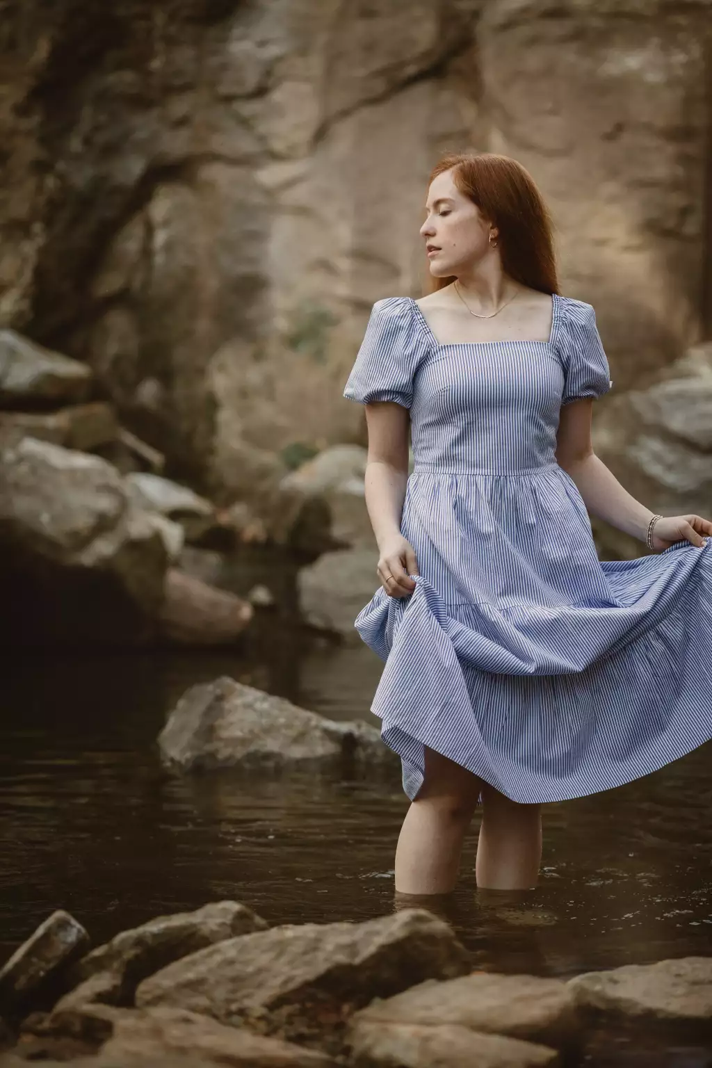 : A young woman with long red hair stands knee-deep in a shallow stream surrounded by large rocks. She wears a short-sleeved, light blue and white striped dress with puffed sleeves and holds the skirt slightly lifted with both hands. She looks to her left with a calm expression, and the background features a rocky, natural setting.