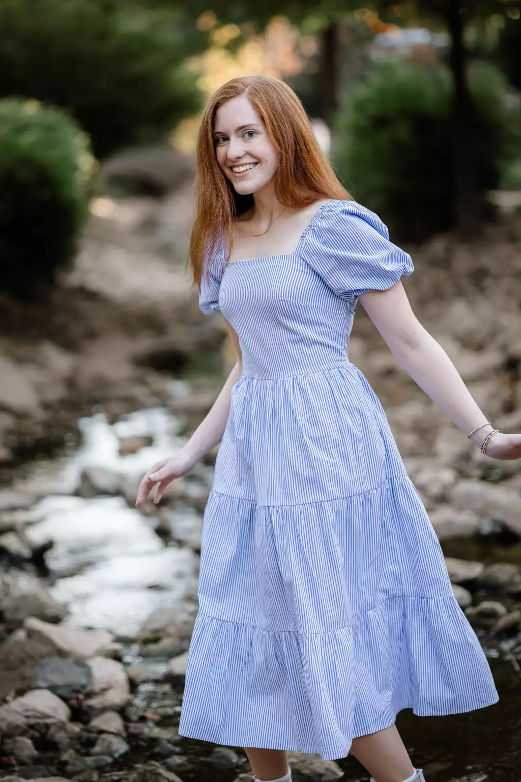 : Young woman with long red hair smiling, wearing a light blue and white striped dress with puffed sleeves, standing outdoors near a rocky stream with blurred greenery in the background.
