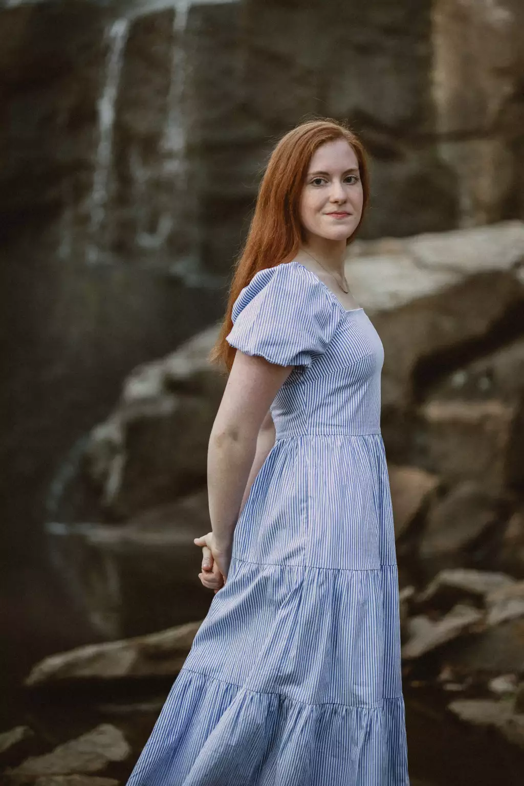 : Young woman with long red hair wearing a light blue and white striped dress standing sideways near rocky terrain with a waterfall in the background, looking at the camera with a slight smile.
