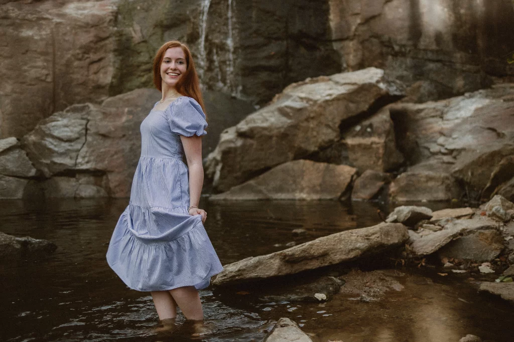 Ryn's senior photos take at the Rock Quaryy Garden, Greenville, SC: Young woman with red hair wearing a blue and white striped dress standing in shallow water by rocks and a small waterfall, smiling and holding the hem of her dress.