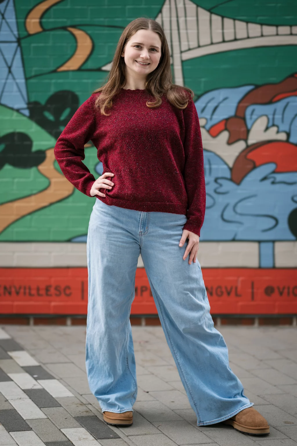 Sabrina's senior photos taken downtown Greenville, SC: Young woman smiling, wearing a red sweater and light blue wide-leg jeans, standing with one hand on her hip in front of a colorful mural on a brick wall.