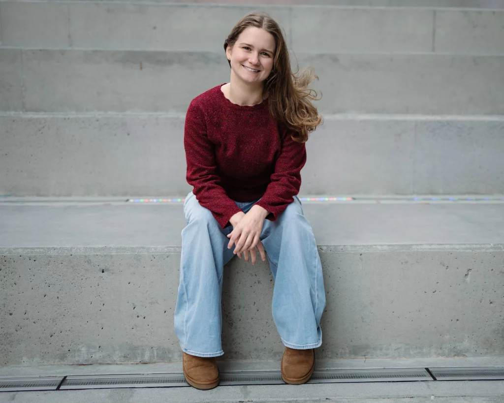 Sabrina's senior photos taken downtown Greenville, SC: Young woman with long hair wearing a red sweater, light blue jeans, and brown boots sitting on concrete steps, smiling at the camera.