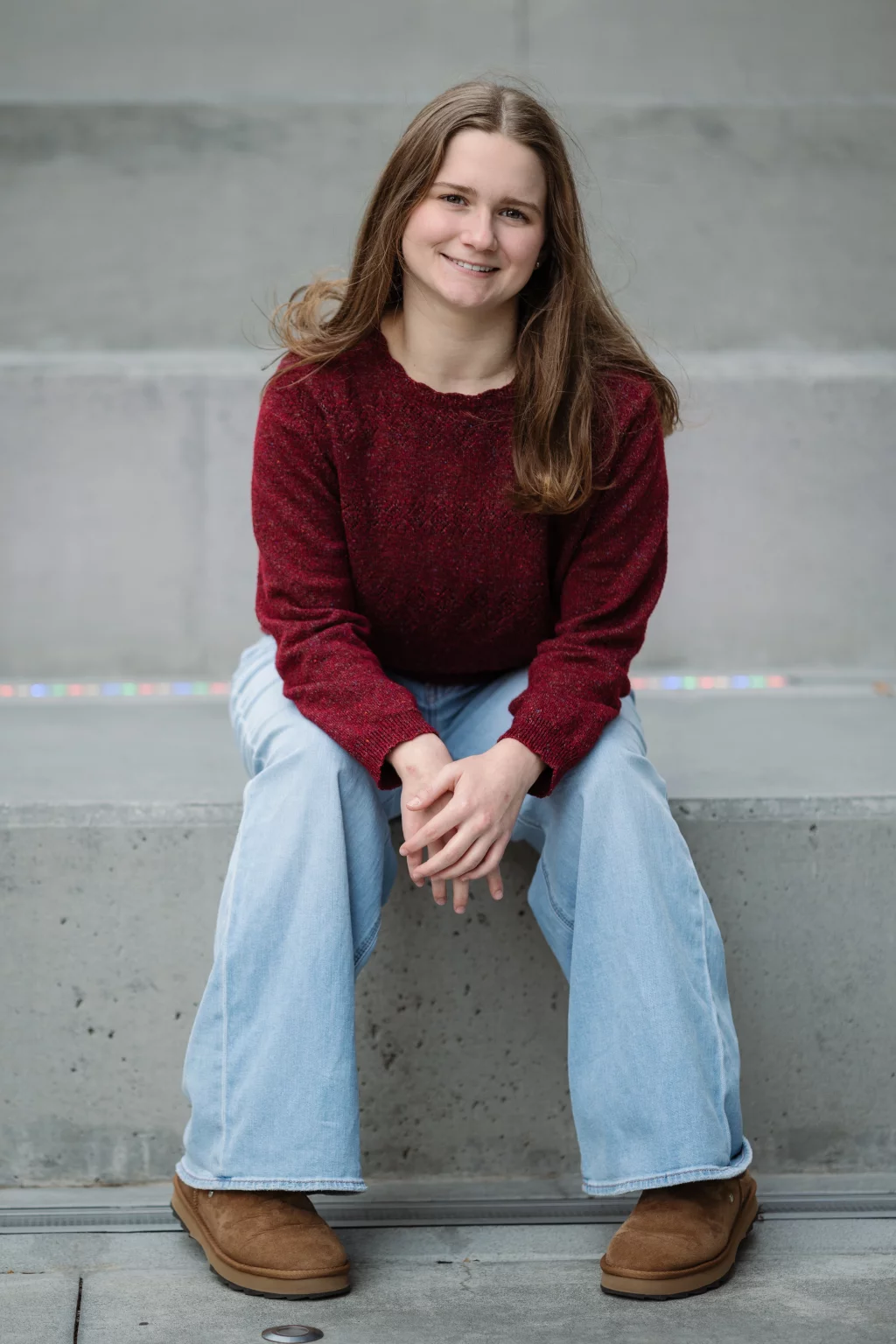 Sabrina's senior photos taken downtown Greenville, SC: Young woman with long brown hair wearing a maroon sweater, light blue jeans, and brown boots, sitting on concrete steps smiling at the camera.