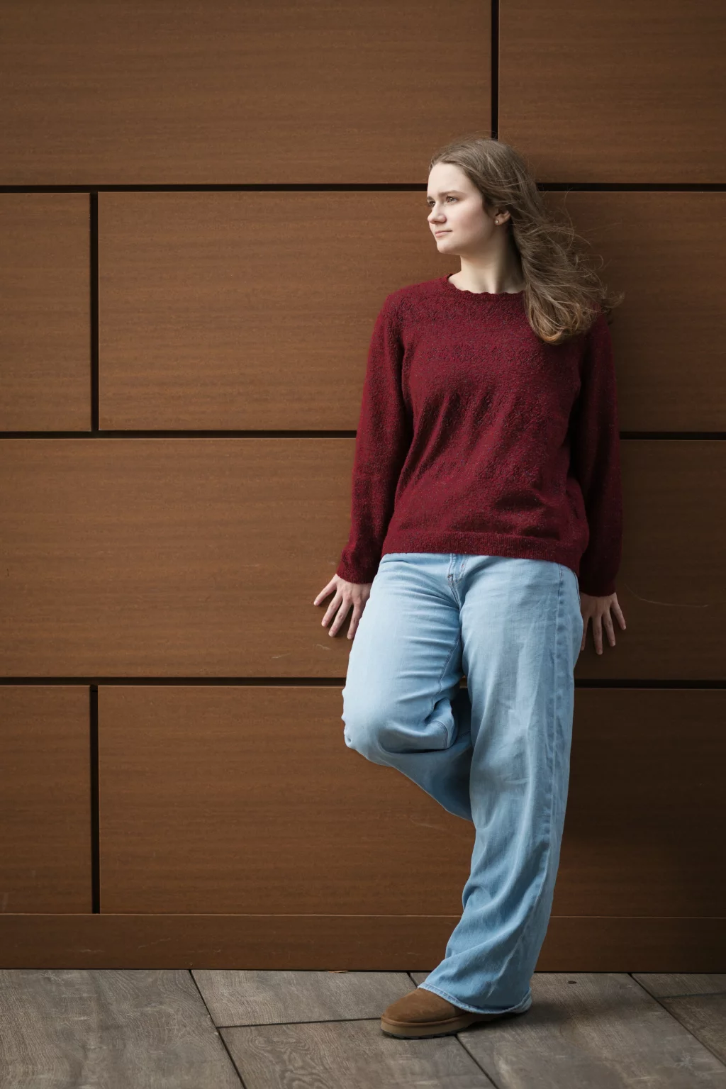 Sabrina's senior photos taken downtown Greenville, SC: Young woman with long hair wearing a red sweater and light blue jeans, leaning against a brown tiled wall with one leg bent and looking to the side.
