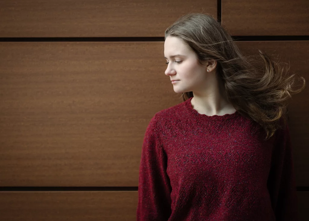 Sabrina's senior photos taken downtown Greenville, SC: Profile of a young woman with light skin and long brown hair blowing in the wind, wearing a textured red sweater, standing against a brown wooden panel background.