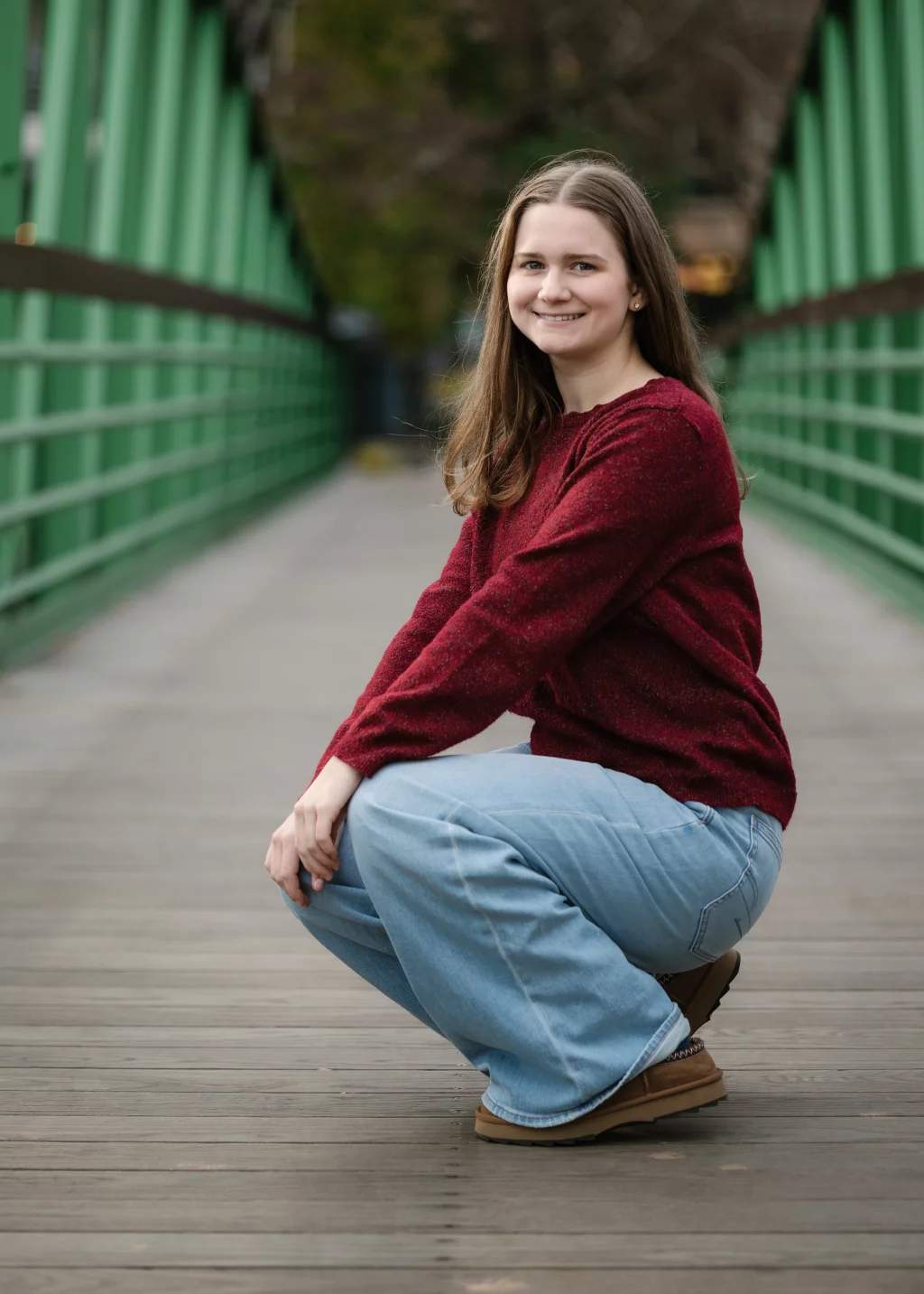 Sabrina's senior photos taken downtown Greenville, SC: Young woman with long brown hair wearing a red sweater, light blue jeans, and brown shoes, squatting and smiling on a wooden bridge with green railings.
