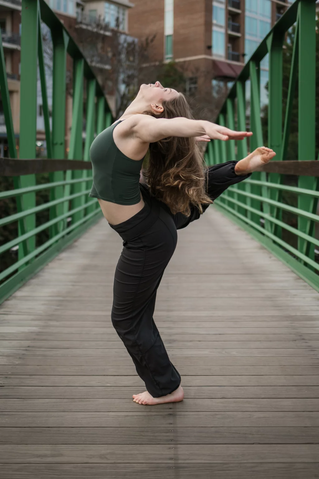 Sabrina's senior photos taken downtown Greenville, SC: Woman in dark green tank top and black pants performing a backbend yoga pose barefoot on a wooden pedestrian bridge with green railings.