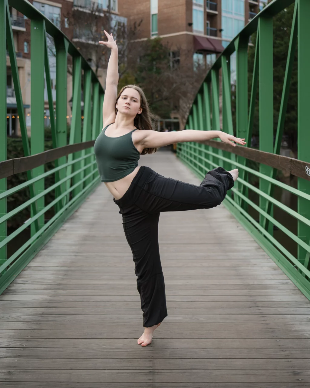 Sabrina's senior photos taken downtown Greenville, SC: Young woman performing a ballet pose on one foot on a green metal pedestrian bridge with wooden planks, urban buildings in the background.