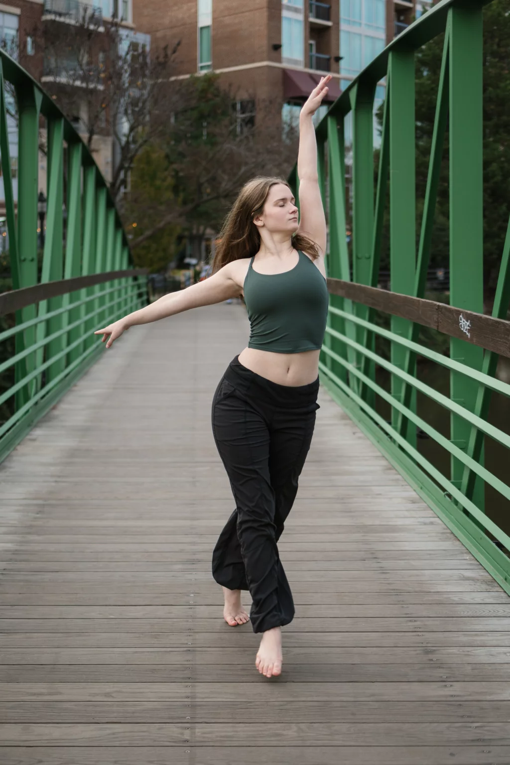 Sabrina's senior photos taken downtown Greenville, SC: Young woman in green tank top and black pants dancing barefoot on a wooden bridge with green railings in an urban setting.