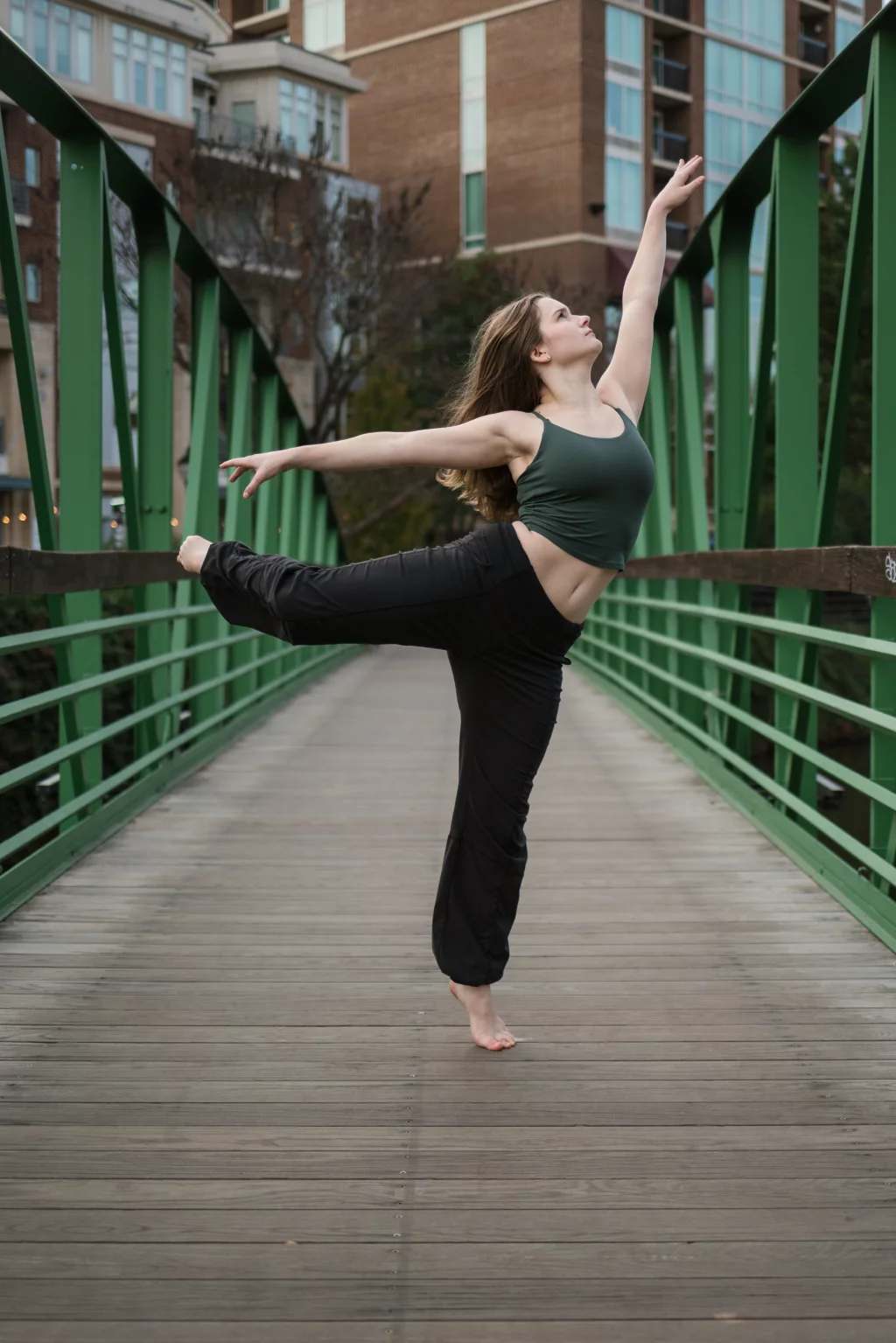 Sabrina's senior photos taken downtown Greenville, SC: Woman in black pants and green tank top balancing on one foot in a dance pose on a green metal pedestrian bridge with buildings in the background.