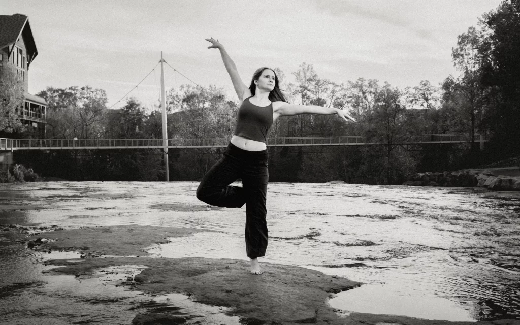 Sabrina's senior photos taken downtown Greenville, SC: Black and white image of a woman balancing barefoot on one leg on a rock in a river, with arms gracefully extended and a bridge and trees in the background.