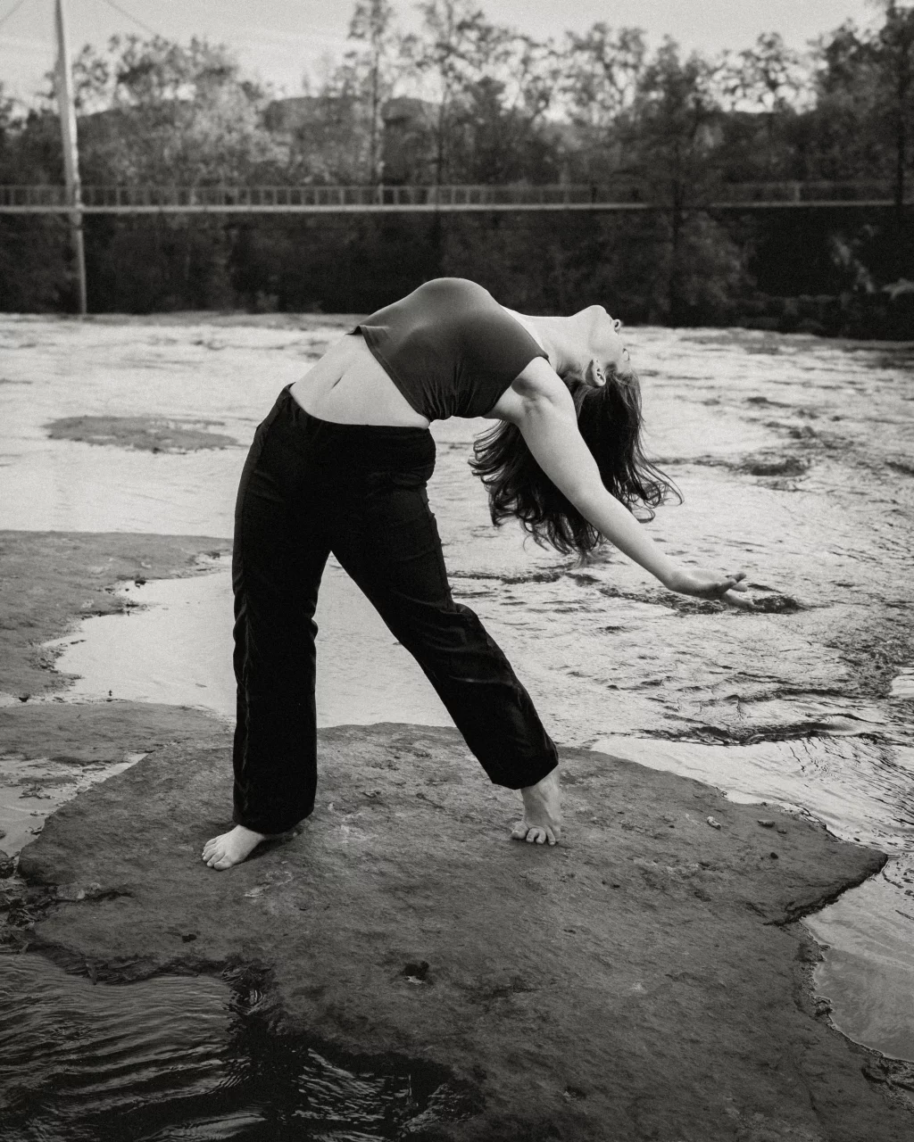 Sabrina's senior photos taken downtown Greenville, SC: Black and white photo of a barefoot woman arching backward on a large flat rock by flowing water, wearing dark loose pants and a cropped top, with trees and a bridge in the background.