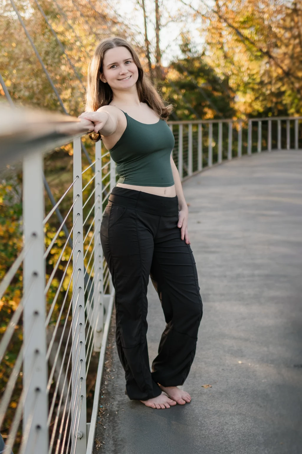 Sabrina's senior photos taken downtown Greenville, SC: Young woman barefoot standing on a curved pedestrian bridge, leaning on the railing, with autumn trees in the background.