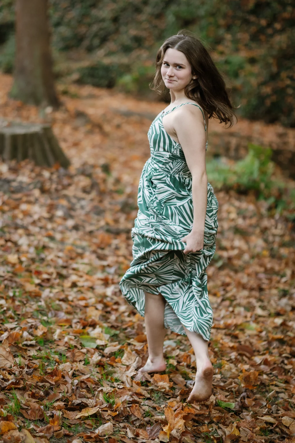 Sabrina's senior photos taken downtown Greenville, SC: Young woman in a green and white leaf-patterned dress walking barefoot through fallen autumn leaves in a forest setting, looking back over her shoulder.