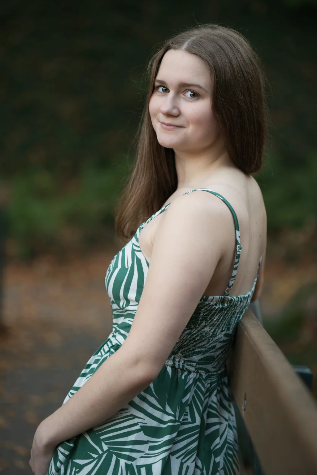 Sabrina's senior photos taken downtown Greenville, SC: Young woman with long brown hair wearing a green and white patterned sleeveless dress, sitting on a wooden bench outdoors, looking over her shoulder and smiling softly.